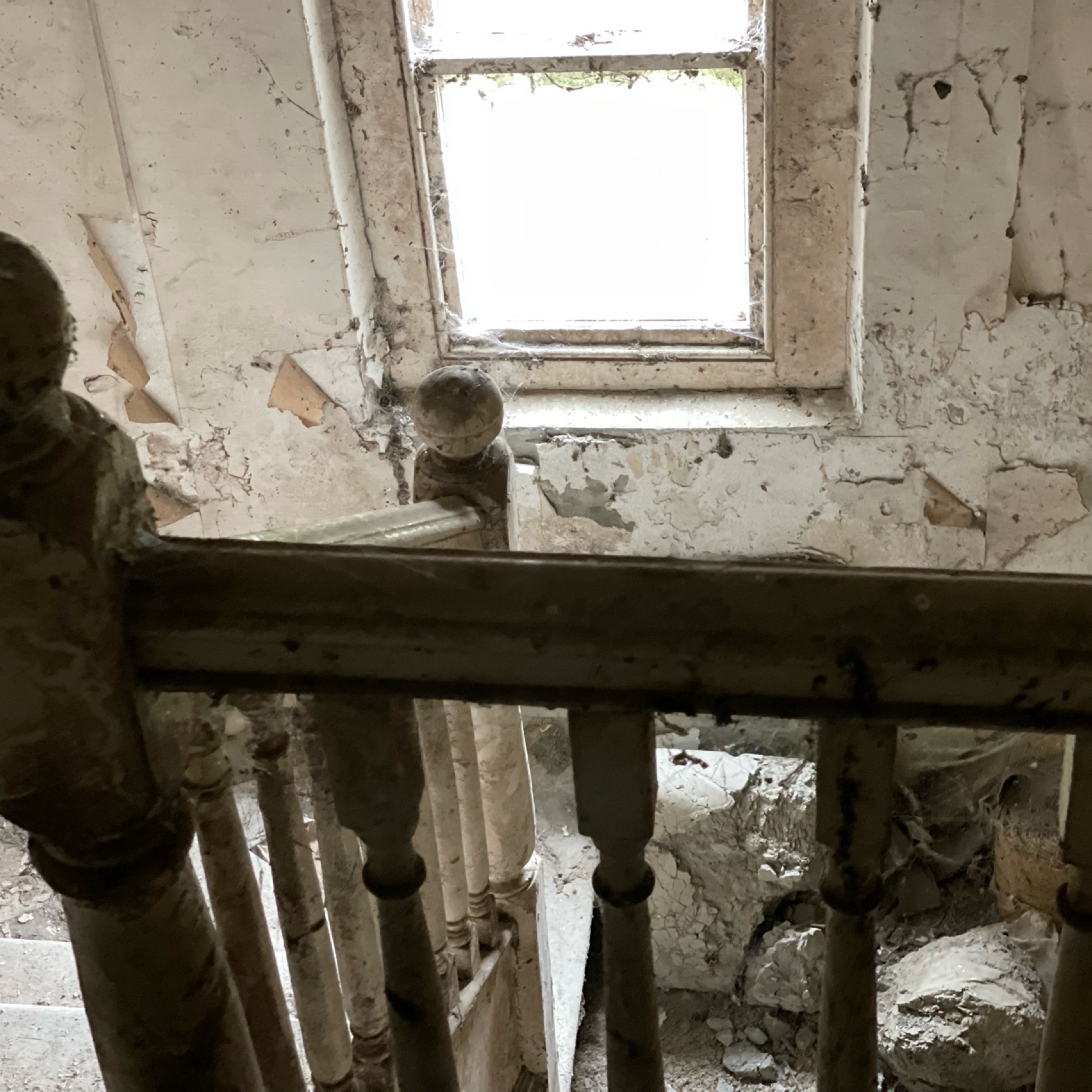 Dilapidated staircase in an abandoned building. Peeling paint, cobwebs, and debris enhance the eerie and neglected atmosphere. Dusty, old sash window provides light.