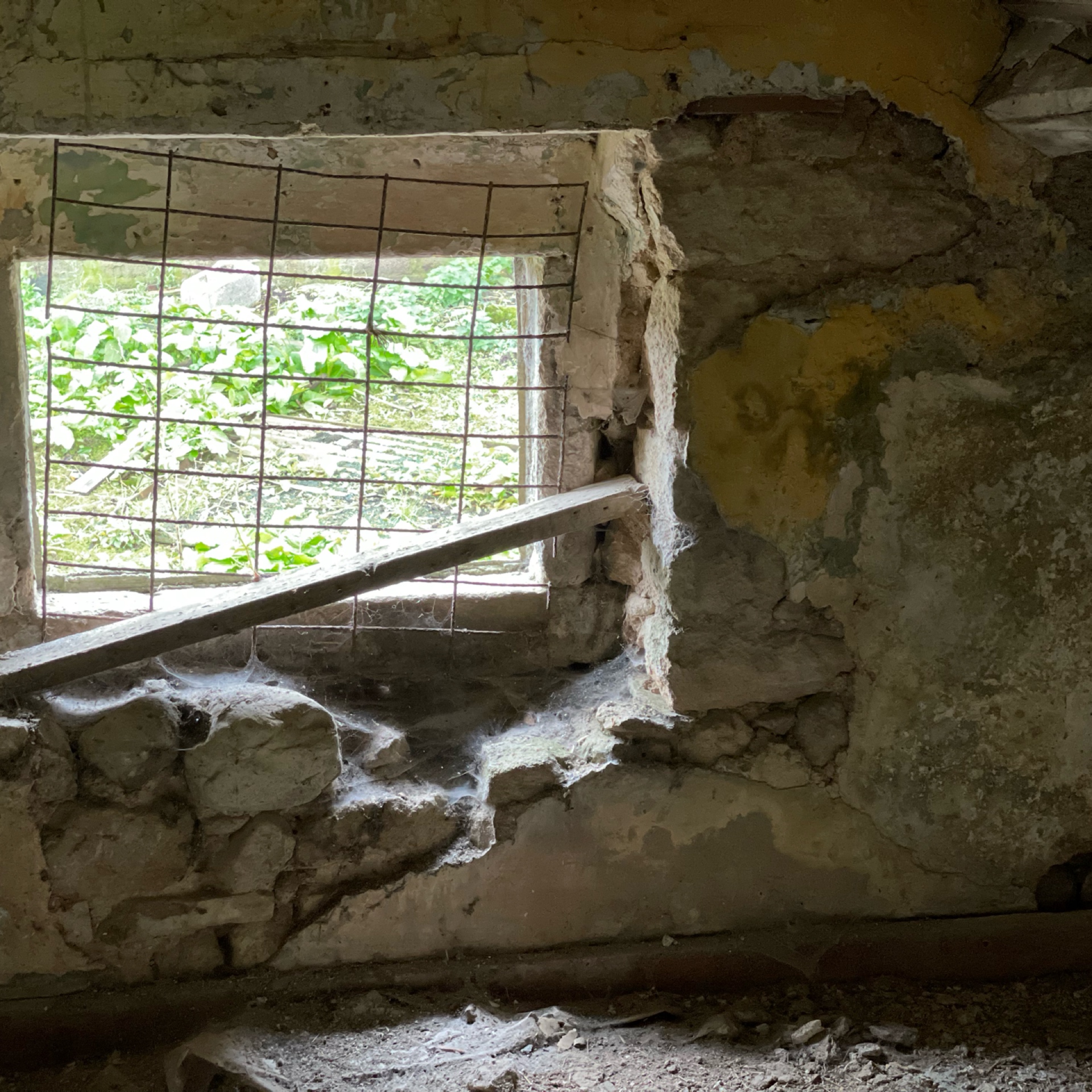 Wire-covered window in stone wall with cobwebs and deteriorating plaster.